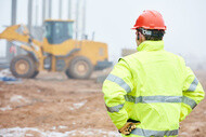 Construction worker in high-visibility clothing and a hard hat with heavy equipment in the background