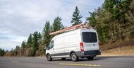 Small compact commercial cargo mini van with ladders on the roof running on the road with trees on the hill to the place of repair or installation work at the request of customers