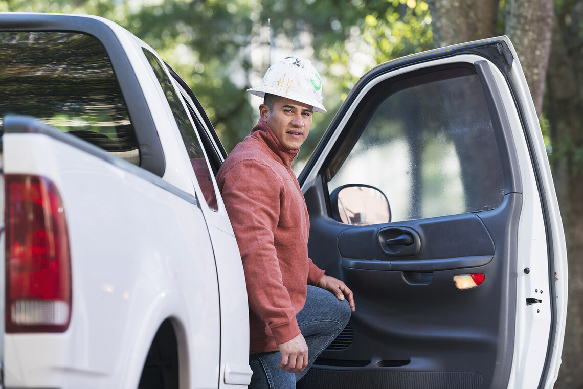 An Hispanic man wearing a worn, white hardhat, getting out of the passenger seat of a white pickup truck.  He is a construction worker wearing a pale orange sweatshirt and jeans, looking at the camera with a serious expression.