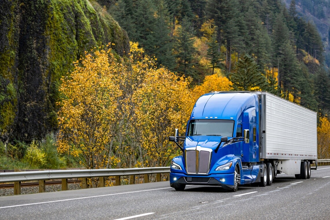 Blue cab semi truck driving with backdrop of mountains and yellow leaves on trees