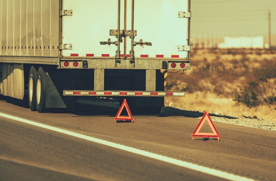 Back of a white semi truck parked on the shoulder of a highway with two emergency triangles behind it