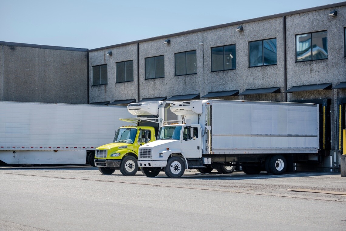 Refrigerated delivery trucks parked at a food and beverage distribution center loading dock, representing fleet operations, route delivery, and cold chain logistics management.