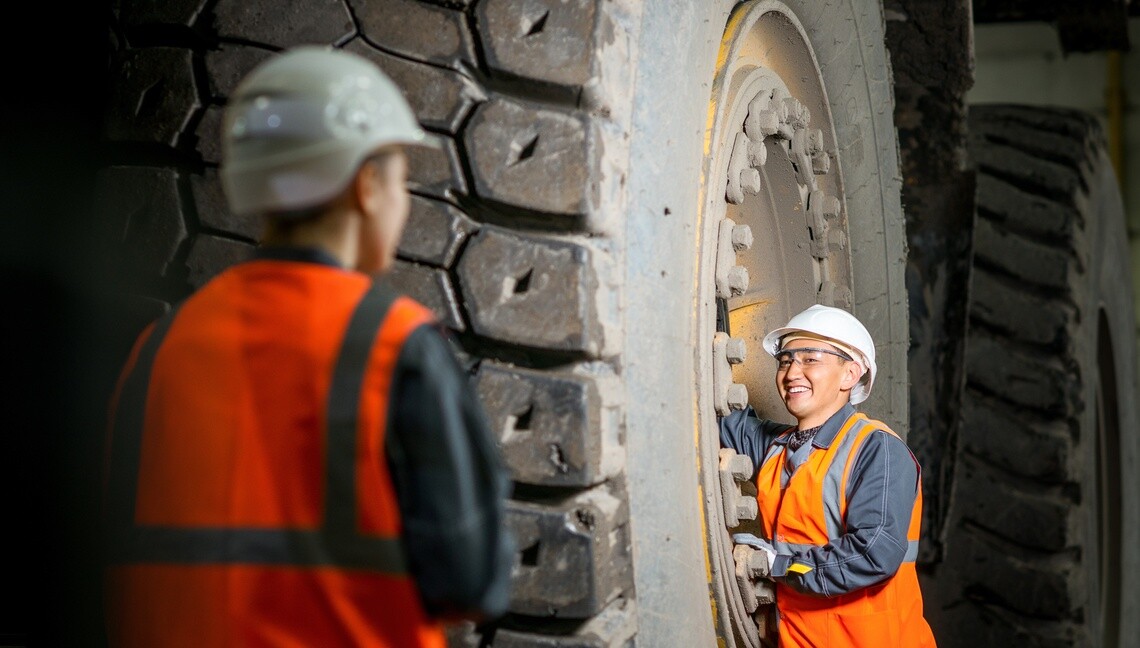 Two workers wearing safety gear beside a large off-road tire.