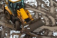 Yellow backhoe loader with large off-road tires operating on muddy, snow-covered terrain.