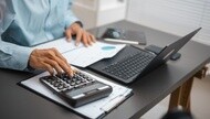 Person using a calculator beside a laptop and financial documents on an office desk.
