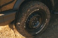 Close-up of a muddy off-road tire on a vehicle parked on dry, grassy terrain. The rugged tire reads "All-terrain T/A" and exudes a rugged, adventurous feel.