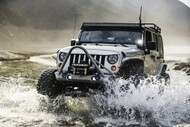 A rugged white Jeep driving through splashing water in a mountainous landscape. Its headlights are on, conveying adventure and excitement.