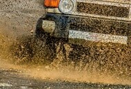 Close-up of an SUV's front end splashing through muddy water, creating a dynamic spray. The scene conveys excitement and off-road adventure.